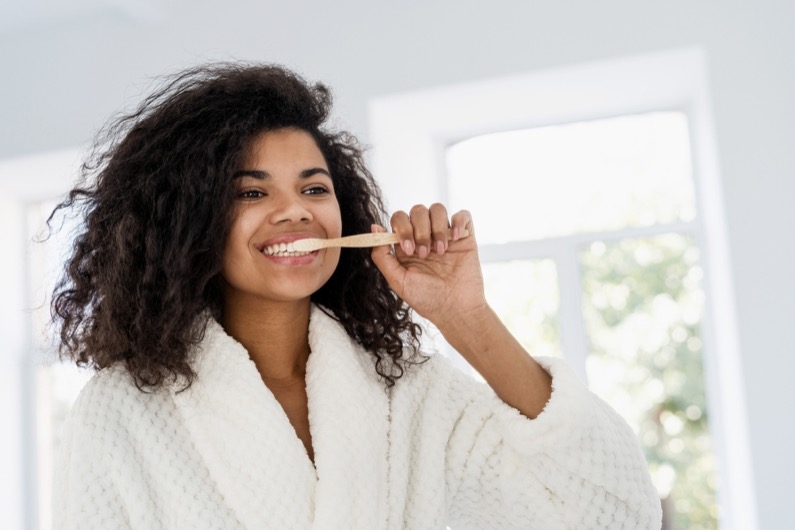 Personal oral hygiene and dental care concept. Portrait of happy young african american woman cleanse sensitive teeth with a bamboo brush and whitening toothpaste. Female in bathrobe looking at mirror