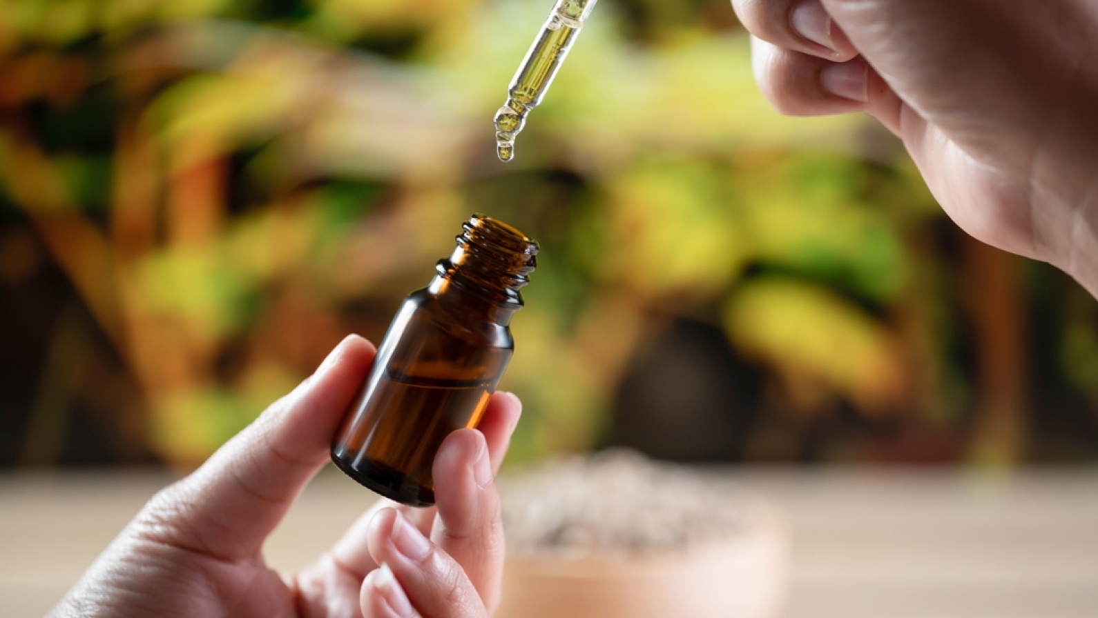 Hands holding a bottle of CBD oil and its dropper lid, with hemp leaf in the background. Legalized CBD product for medical purposes.