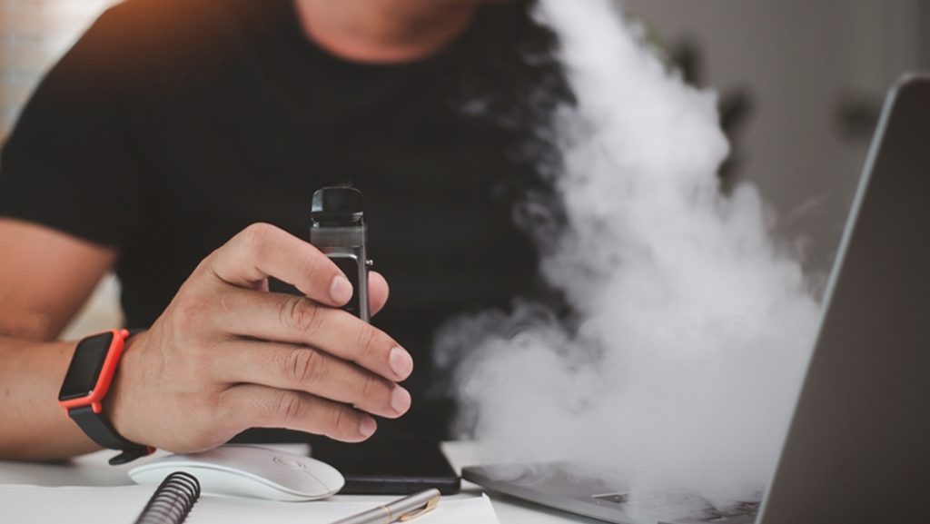 young man using disposable electronic cigarette at table in office