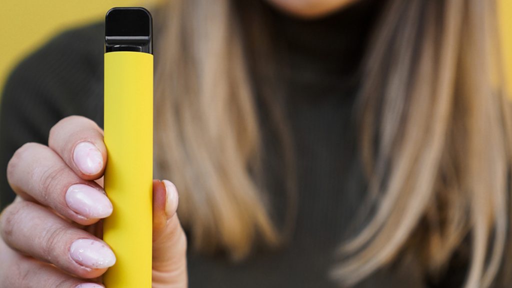 Yellow disposable electronic cigarette in female hand. Bright yellow background