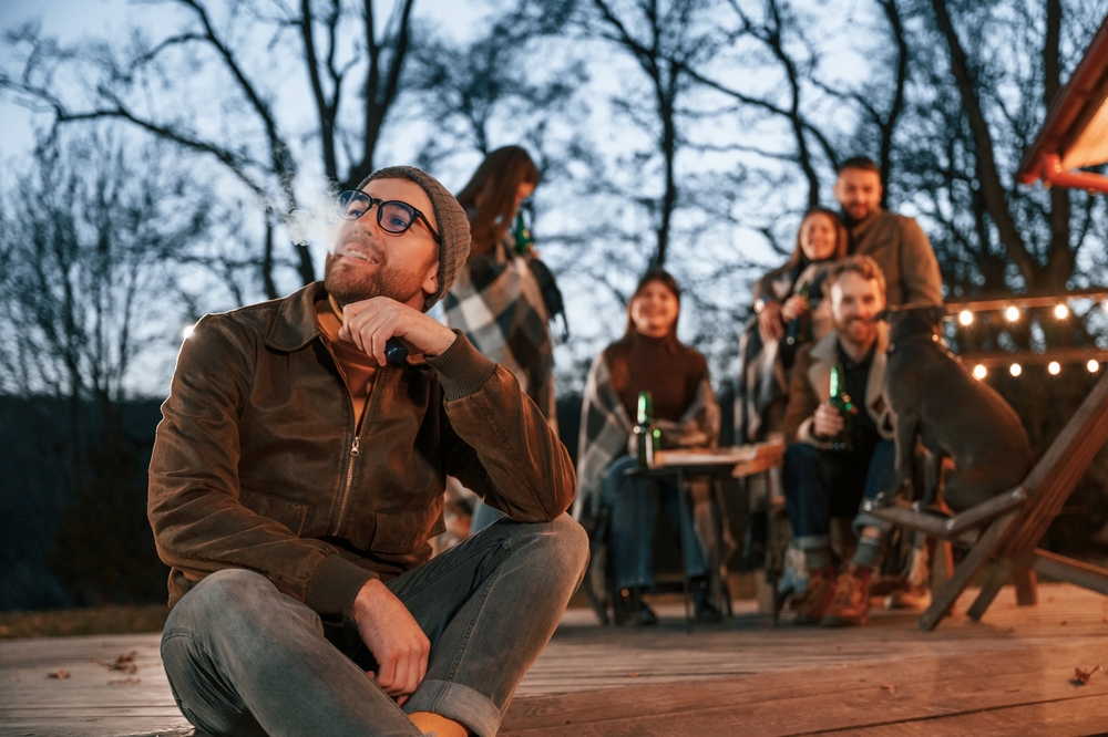 Man is smoking. Group of people is spending time together on the backyard at evening time.